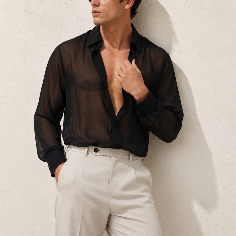 May include: A black long-sleeved shirt made of a sheer, translucent fabric featuring a classic collar, a button-down front, and buttoned cuffs. The shirt is worn unbuttoned at the top and is paired with light beige trousers that have a visible button closure.