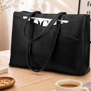 May include: A black rectangular tote bag with two thin shoulder straps and a V-shaped front pocket detail. The bag features gold-toned hardware connecting the straps to the main body, which has a smooth finish and a structured, upright design.