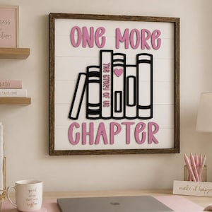 May include: A square wall sign with a dark wood frame and white shiplap background. It features pink 3D lettering that reads ONE MORE CHAPTER and a black graphic of five books, one of which is labeled THE STORY OF US with a small pink heart.