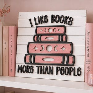 May include: A square white wooden sign with horizontal slats featuring black 3D lettering that reads I LIKE BOOKS MORE THAN PEOPLE. In the centre, three stacked pink wooden books are displayed, each decorated with black moon phase cutouts.