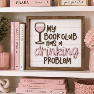 May include: A square wooden sign with a white shiplap background and a dark brown frame. The sign features a pink wine glass icon and the text "MY BOOK CLUB HAS A drinking PROBLEM" in a mix of dark brown and pink raised 3D lettering.