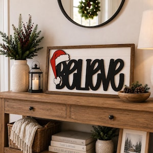 Op de afbeelding: Een rechthoekig houten bord met een witte achtergrond in latjesstijl en een donkerbruine lijst. Het woord BELIEVE is geschreven in grote, zwarte cursieve letters, met een rood-witte kerstmuts die op de letter B rust.
