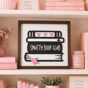 May include: A square wooden sign with a dark brown frame and a white shiplap background. The sign features a black layered cutout of a stack of three books with the text "SMUTTY BOOK CLUB" in white, three small pink hearts, and a pink bookmark.