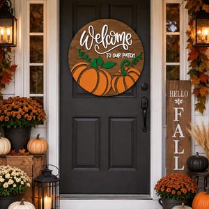 May include: A round wooden door sign featuring the white text Welcome to our patch above two orange pumpkins with green stems and vines. The sign is displayed on a black door surrounded by fall decor including orange mums and pumpkins.