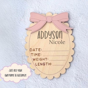 Pode incluir: Um letreiro de anúncio de bebé em madeira com um laço rosa no topo. O letreiro tem o nome "Addyson Nicole" e espaços para data, hora, peso e comprimento. O texto "Just add your own name to customize" também está presente.