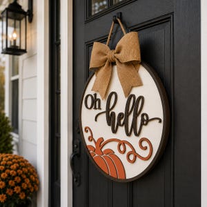 May include: A circular wooden door sign featuring a cream-colored background with the dark brown text Oh hello. The sign includes a layered orange pumpkin graphic with swirling vines at the bottom and a large tan burlap bow attached at the top.