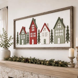 May include: A rectangular wall art piece featuring four stylized houses and several evergreen trees in red, white, and green, enclosed in a rustic brown wooden frame. The houses have cut-out windows and doors, arranged against a white background.