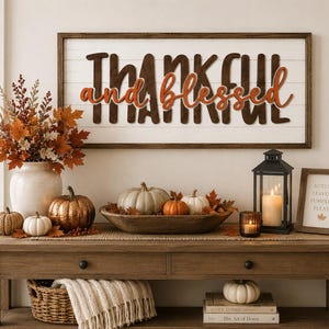 Pode incluir: Uma placa de parede retangular com fundo de ripas brancas e moldura de madeira escura, apresentando o texto Thankful em letras de bloco castanho-escuras e as palavras and blessed em escrita cursiva laranja, exibida sobre uma mesa consola de madeira.