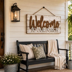 Peut inclure: Un panneau rectangulaire en bois avec une finition blanche vieillie, arborant le texte Welcome en lettres cursives marron en relief et TO OUR PORCH en lettres capitales blanches. Le panneau est suspendu par une corde marron.