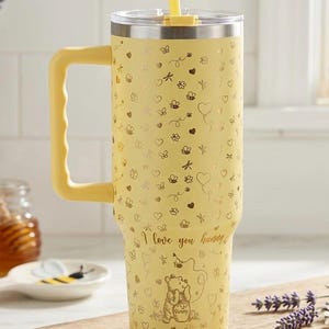 May include: Yellow insulated tumbler with a handle, straw, and lid. The tumbler is decorated with gold hearts, bees, and the phrase "I love you hunny" with a Winnie the Pooh graphic. A charming and functional drinkware item.