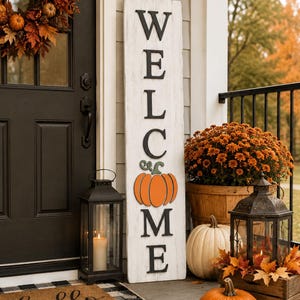 Può includere: Un alto cartello rettangolare in legno bianco con finitura anticata, caratterizzato dalla scritta verticale nera WELCOME, in cui la lettera O è sostituita da un'illustrazione di una zucca arancione brillante. Il cartello è accanto a una lanterna.