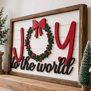 May include: A rectangular wooden sign with a white shiplap background and a dark brown frame. The sign features the red text JOY with a green wreath and red bow replacing the letter O, and the black cursive text to the world below it.