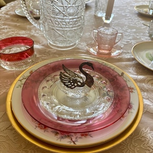 May include: A clear glass pitcher with a starburst pattern, a small pink glass cup with two handles, a red glass bowl, and a stack of plates featuring a metal swan figurine on top. The swan has a dark finish and detailed wing cutouts.