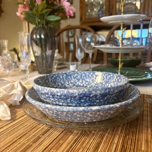 May include: A dining table setting featuring a stack of blue and white speckled bowls atop a clear glass plate. The bowls are on a bamboo placemat. A vase of pink flowers and glassware are in the background.