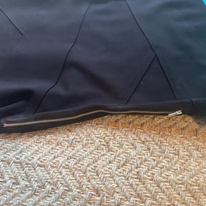May include: Close-up of a black garment with a silver zipper. The garment has a geometric design with angled seams. A turquoise fabric is visible on the right side of the image. The garment is lying on a textured, beige surface.