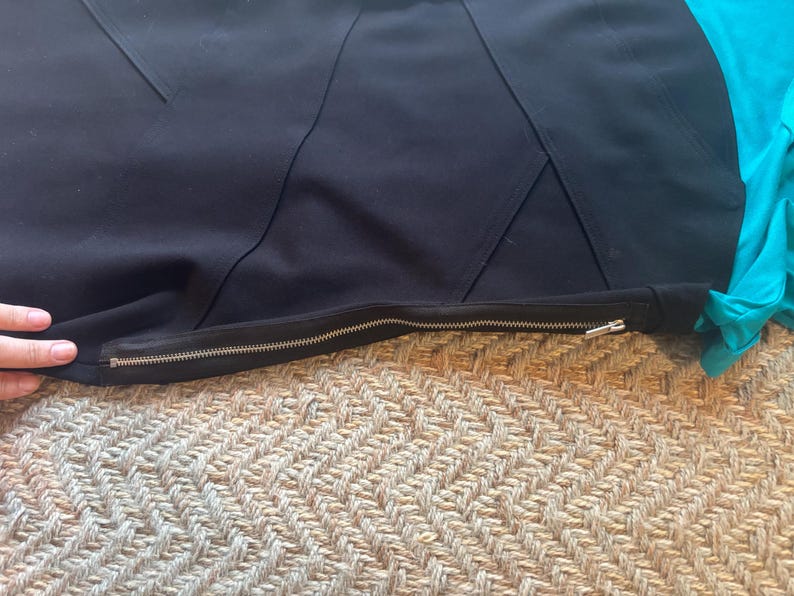 May include: Close-up of a black garment with a silver zipper. The garment has a geometric design with angled seams. A turquoise fabric is visible on the right side of the image. The garment is lying on a textured, beige surface.