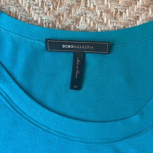 May include: A close-up of a teal-colored garment with a round neckline. The label reads "BCBGMAXAZRIA" in a rectangular black tag. Below the tag is a smaller tag that reads "Made in China" and the number "06".