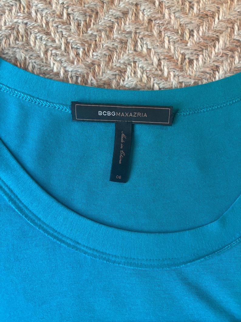 May include: A close-up of a teal-colored garment with a round neckline. The label reads "BCBGMAXAZRIA" in a rectangular black tag. Below the tag is a smaller tag that reads "Made in China" and the number "06".