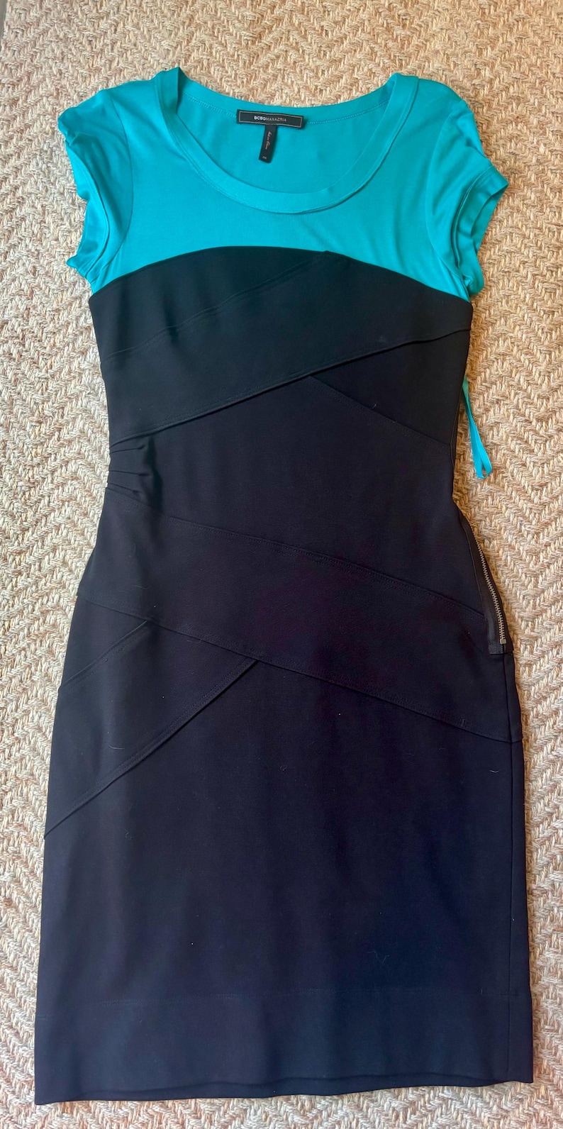 May include: A two-tone dress with a turquoise top and a black skirt. The dress features a fitted silhouette with a zipper closure and diagonal seam detailing. The dress is made of a smooth fabric.
