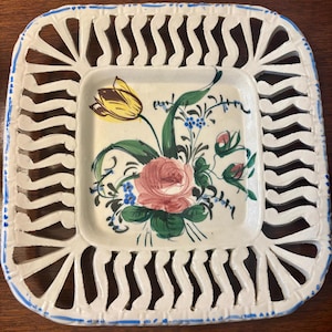 May include: A square off-white ceramic plate with a lattice-style openwork border accented by thin blue painted lines. The center features a hand-painted floral arrangement including a yellow tulip, a pink rose, and small blue flowers with green leaves.