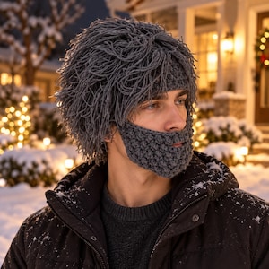 May include: A dark gray crocheted hat featuring a thick, shaggy fringe texture that mimics hair, paired with an integrated, textured crocheted beard covering the chin and jaw area. The accessory is crafted from dark gray yarn in a dense stitch.