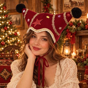 May include: A deep red velvet jester-style hat featuring two pointed peaks topped with black pom-poms. The hat is decorated with gold braided trim, small white pearl-like beads along the edge, and various colourful glittery star-shaped appliqués.