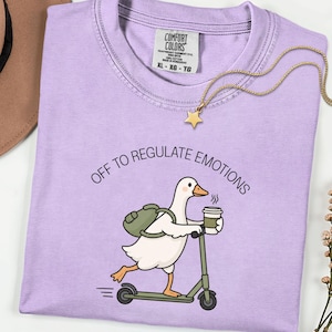 Puede incluir: Una camiseta de color morado claro con el gráfico de un ganso blanco con pico y patas naranjas, que lleva una mochila verde, monta un patinete verde y sostiene una taza de café. El texto dice OFF TO REGULATE EMOTIONS en letras negras.