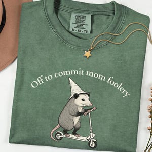 Peut inclure: Un t-shirt vert sauge avec une illustration d'un opossum portant un chapeau pointu et montant une trottinette blanche. Le texte "Off to commit mom foolery" est imprimé en blanc au-dessus de l'opossum. Le t-shirt porte une étiquette Comfort Colors.