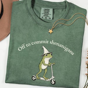 May include: A sage green t-shirt featuring a graphic of a frog wearing a pointed hat and riding a white scooter. The text above the frog reads, Off to commit shenanigans. The shirt has a Comfort Colors label at the neck and a gold star necklace.