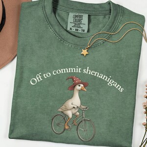 May include: A sage green crewneck t-shirt featuring a graphic of a white goose wearing a red wizard hat and riding a bicycle. The text "Off to commit shenanigans" is printed in white above the goose. A gold star pendant necklace rests on the collar.