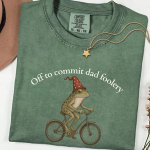 Puede incluir: Una camiseta verde salvia con el gráfico de una rana con un sombrero de mago rojo con estrellas montada en bicicleta. El texto "Off to commit dad foolery" está impreso en blanco sobre la rana. Un collar con colgante de estrella dorada descansa sobre ella.
