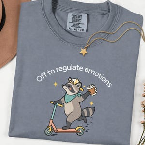 Op de afbeelding: Een leiblauw t-shirt met een afbeelding van een wasbeer met een gele pet en een groenblauwe bandana, rijdend op een step met een koffiebeker. De tekst erboven luidt, Off to regulate emotions. Een gouden sterrenketting ligt op de kraag.