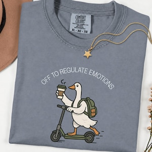 May include: A slate blue t-shirt featuring a graphic of a white goose riding a scooter while holding a coffee cup and wearing a green backpack. The text above the goose reads "OFF TO REGULATE EMOTIONS" in white. The shirt has a Comfort Colors label.