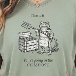 Op de afbeelding: Een saliegroen t-shirt met een zwart-witte lijntekening van een kikker in een schort die een riek vasthoudt naast een houten compostbak. De tekst luidt: That's it. You're going in the COMPOST.