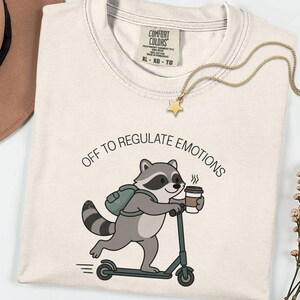 Può includere: Una t-shirt color crema con la grafica di un procione su un monopattino, che indossa uno zaino e tiene in mano una tazza di caffè. Il testo sopra il procione recita OFF TO REGULATE EMOTIONS. La t-shirt presenta un'etichetta del marchio Comfort Colors.
