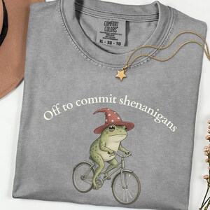 May include: A grey t-shirt featuring a graphic of a green frog wearing a red wizard hat with white stars, riding a bicycle. The text "Off to commit shenanigans" is printed in white above the frog. A gold star pendant necklace rests on the shirt.