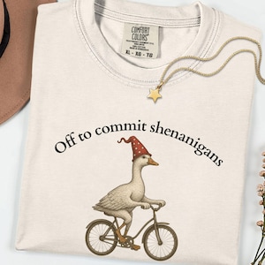 May include: A cream-coloured crewneck t-shirt featuring a graphic of a goose wearing a red wizard hat with stars, riding a bicycle. The text "Off to commit shenanigans" is printed in black above the goose. The shirt has a Comfort Colors brand label.