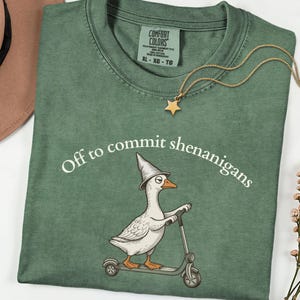May include: A sage green crewneck t-shirt featuring a graphic of a white goose wearing a pointed hat and riding a scooter. The text "Off to commit shenanigans" is printed in white above the goose. The shirt has a Comfort Colors brand label.