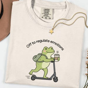 May include: A cream-coloured t-shirt featuring a graphic of a green frog with a backpack riding a scooter while holding a coffee cup. The text above the frog reads, Off to regulate emotions. The shirt has a Comfort Colors brand label in the neck.
