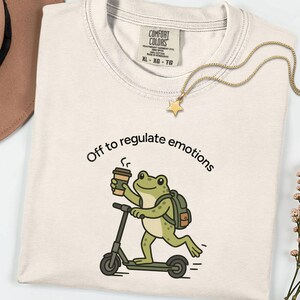Puede incluir: Una camiseta de color crema con un gráfico de una rana verde montada en un patinete mientras sostiene una taza de café. El texto sobre la rana dice "Off to regulate emotions". La camiseta tiene una etiqueta de la marca Comfort Colors en el cuello.