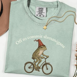 May include: A light green t-shirt featuring a graphic of a frog wearing a red star-patterned wizard hat while riding a bicycle. The text "Off to commit shenanigans" is printed in white above the frog. A gold star pendant necklace rests on the collar.