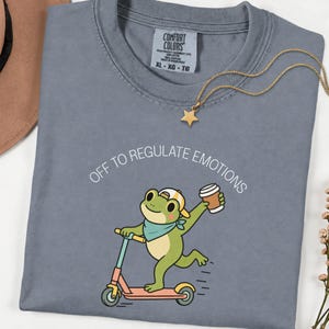 Peut inclure: Un t-shirt bleu ardoise avec un motif représentant une grenouille verte portant une casquette jaune et un foulard bleu, sur une trottinette avec un gobelet de café. Le texte au-dessus indique OFF TO REGULATE EMOTIONS en lettres blanches.