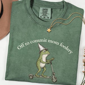 Könnte beinhalten: Ein salbeigrünes T-Shirt mit einer Grafik eines Frosches, der einen spitzen Hut trägt und auf einem Roller fährt. Der Text "Off to commit mom foolery" ist in Weiß darüber gedruckt. Das Comfort Colors Etikett zeigt die Größe XL an.