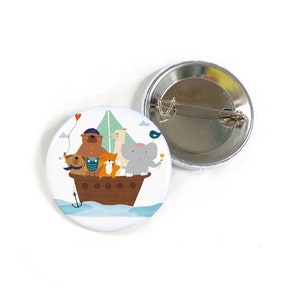 Puede incluir: Un botón blanco con una ilustración colorida de un barco que lleva un oso, una llama, un búho, un zorro y un elefante. El barco navega en aguas azules con un globo rojo en forma de corazón en el cielo.