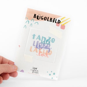 Può includere: Un foglio di trasferimento termoadesivo bianco con il testo "Tanze wild, liebe lebendig, lache laut" in lettere colorate. Il foglio è confezionato in una busta bianca con un design rosa e giallo.