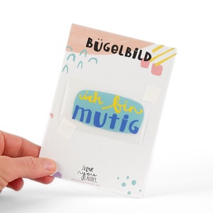 Könnte beinhalten: Eine weiße Karte mit blauen, gelben und rosa abstrakten Formen. Die Karte sagt "Bügelbild" und "ich bin mutig". Die Karte ist für einen Bügelbild-Aufkleber.
