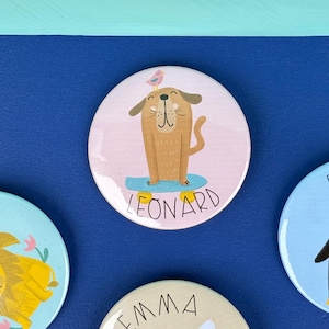 Pode incluir: Um crachá redondo com fundo rosa claro apresentando um cão castanho em cima de um skate azul. Um pequeno pássaro cor-de-rosa está pousado na cabeça do cão. O nome LEONARD está impresso em letras maiúsculas pretas sob o skate.