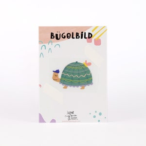 Op de afbeelding: Een strijkplaatje met een cartoon schildpad die een blauwe hoed draagt. Het strijkplaatje is groen en geel en bevindt zich op een witte kaart met de tekst "Bügelbild" in zwart.
