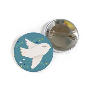 Könnte beinhalten: Ein blauer Knopf mit einer weißen Taube mit geschlossenen Augen, die mit einem Blumenmuster fliegt. Der Knopf hat eine silberne Stecknadel auf der Rückseite.