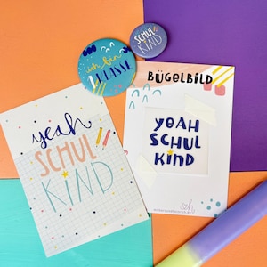Könnte beinhalten: Zwei runde Ansteckbuttons, einer blau mit dem Text "ich bin 1. KLASSE" und einer lila mit "SCHUL KIND", sowie zwei rechteckige Karten. Eine Karte zeigt den Text "yeah SCHUL KIND" und Karomuster, die andere "BÜGELBILD" und "yeah SCHUL KIND".